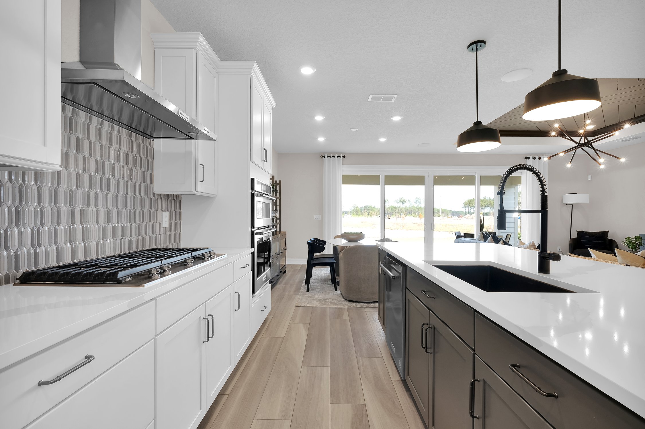 Pulte Homes Wildlight open concept kitchen and living room. White cabinets on the left side of the kitchen with gray backsplash. Large island on the right with dark cabinets and white counters.