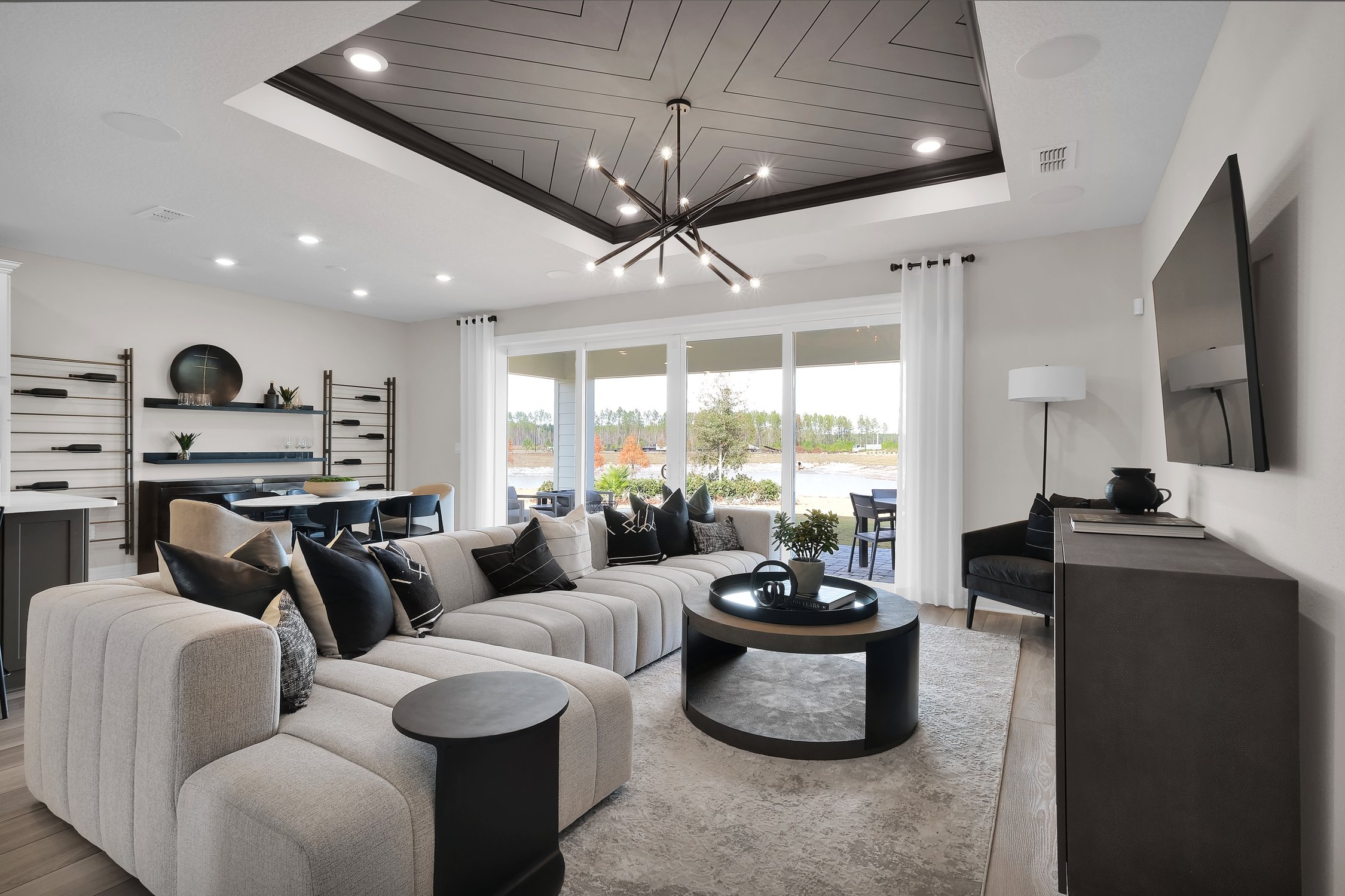 Pulte Homes Wildlight large living room with light gray sectional, wall-mounted tv, recessed ceiling with dark shiplap accents and a wall of doors that open to the patio.