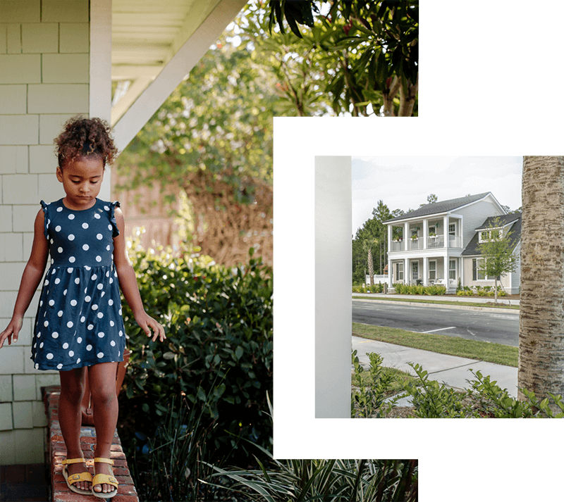 African American girl wearing a navy and white polka dot dress and yellow sandals, walking along a front stoop of a house. There are shrubs and trees in the background. White two-story home with four columns going all the way up to the second floor. The home has a large second story balcony. 