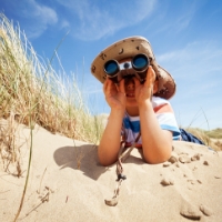 Little boy with a sun hat laying in the sand on his elbows looking into binoculars. The sky is blue and there is beach grass next to him at Big Talbot Island State Park.