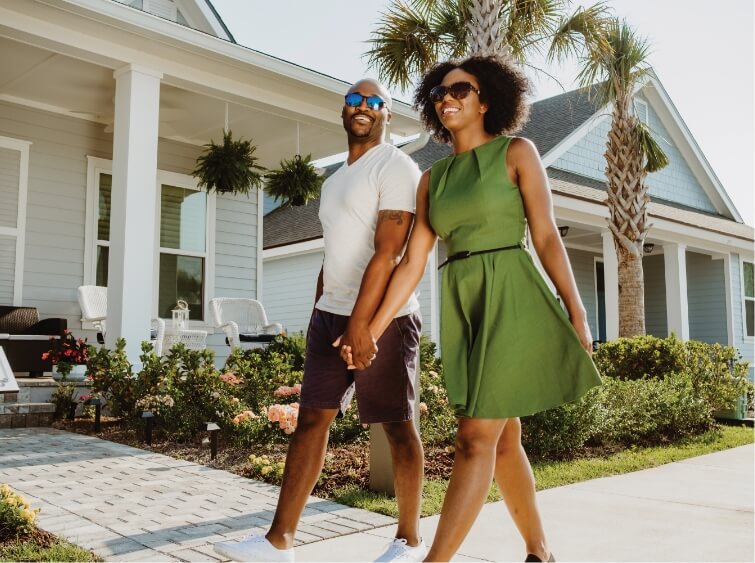 African American couple walking down a street in Wildlight lined with houses, holding hands. They are both wearing sunglasses and the woman is wearing a green dress.