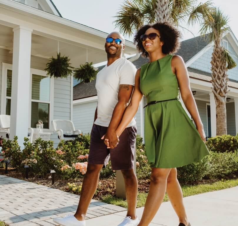 African American couple walking down a street lined with houses, holding hands. They are both wearing sunglasses and the woman is wearing a green dress.