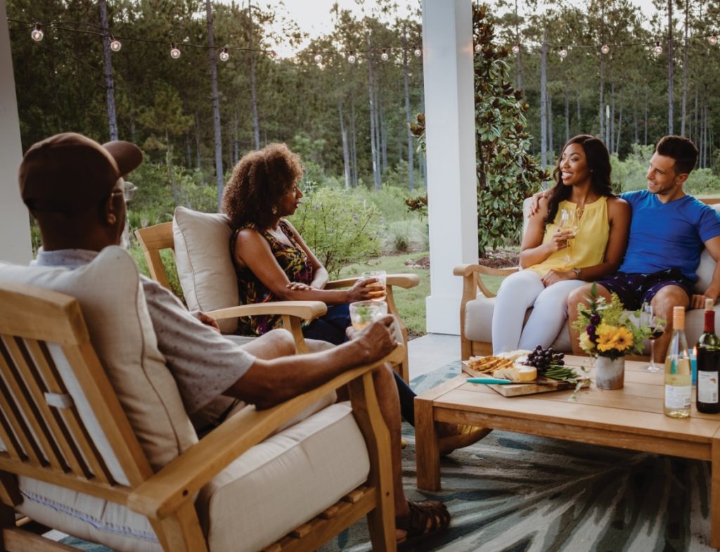 Four adults sitting on a back porch with coffee table with wine and cheese board. One couple is sitting on a couch and the other is sitting on two chairs. They are surrounded by woods and grass.