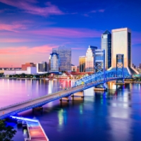 A wide shot of downtown Jacksonville with the sun setting and the tall buildings reflecting off the water in front of them.
