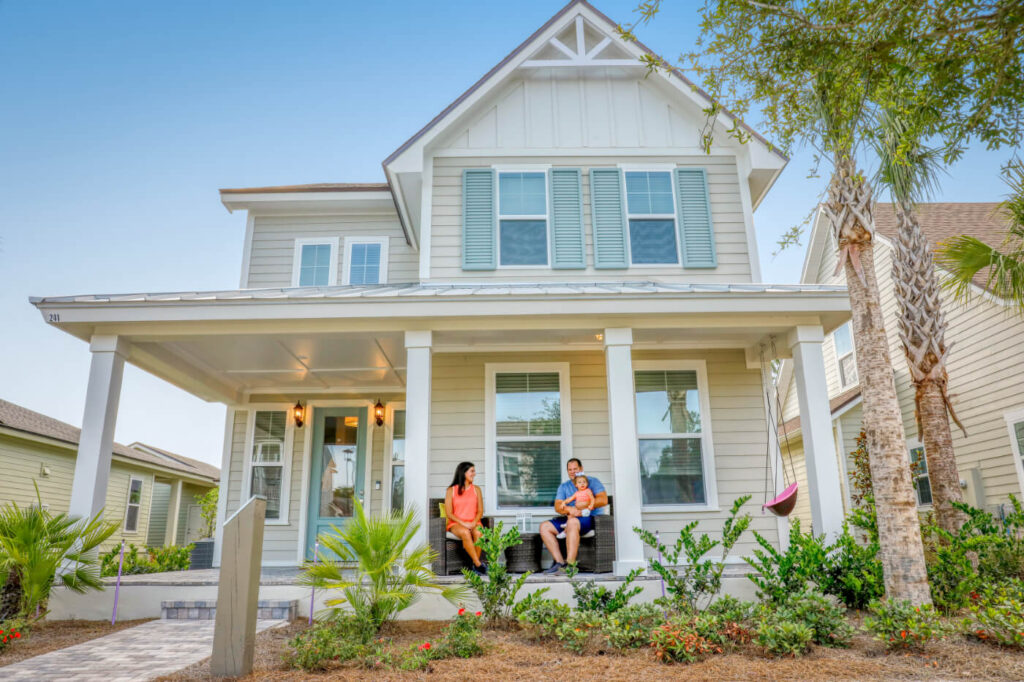 Tan house with pale green shutters with a man, woman and baby sitting on the front porch.