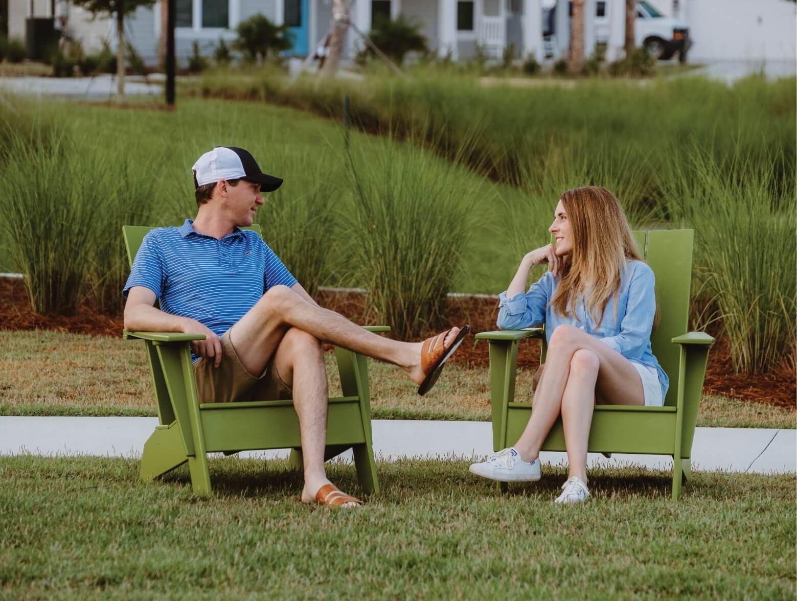 A man and a woman sitting on green lawn chairs in a park chatting. The man is wearing a black and white hat and a blue and white striped shirt. The woman is wearing a blue blouse.