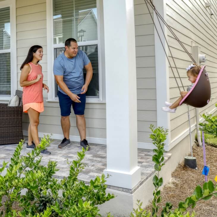 A man and woman pushing a baby on a swing on a front porch of a white, two-story home.