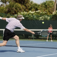 A man and woman playing 1v1 tennis at Amelia National Tennis Club. The man is mid-swing.