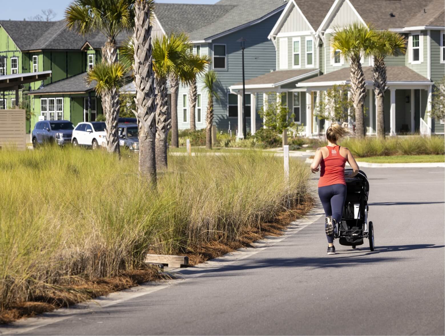 Woman in a red tank top going for a run in the neighborhood with a stroller on a trail near a street lined with houses.