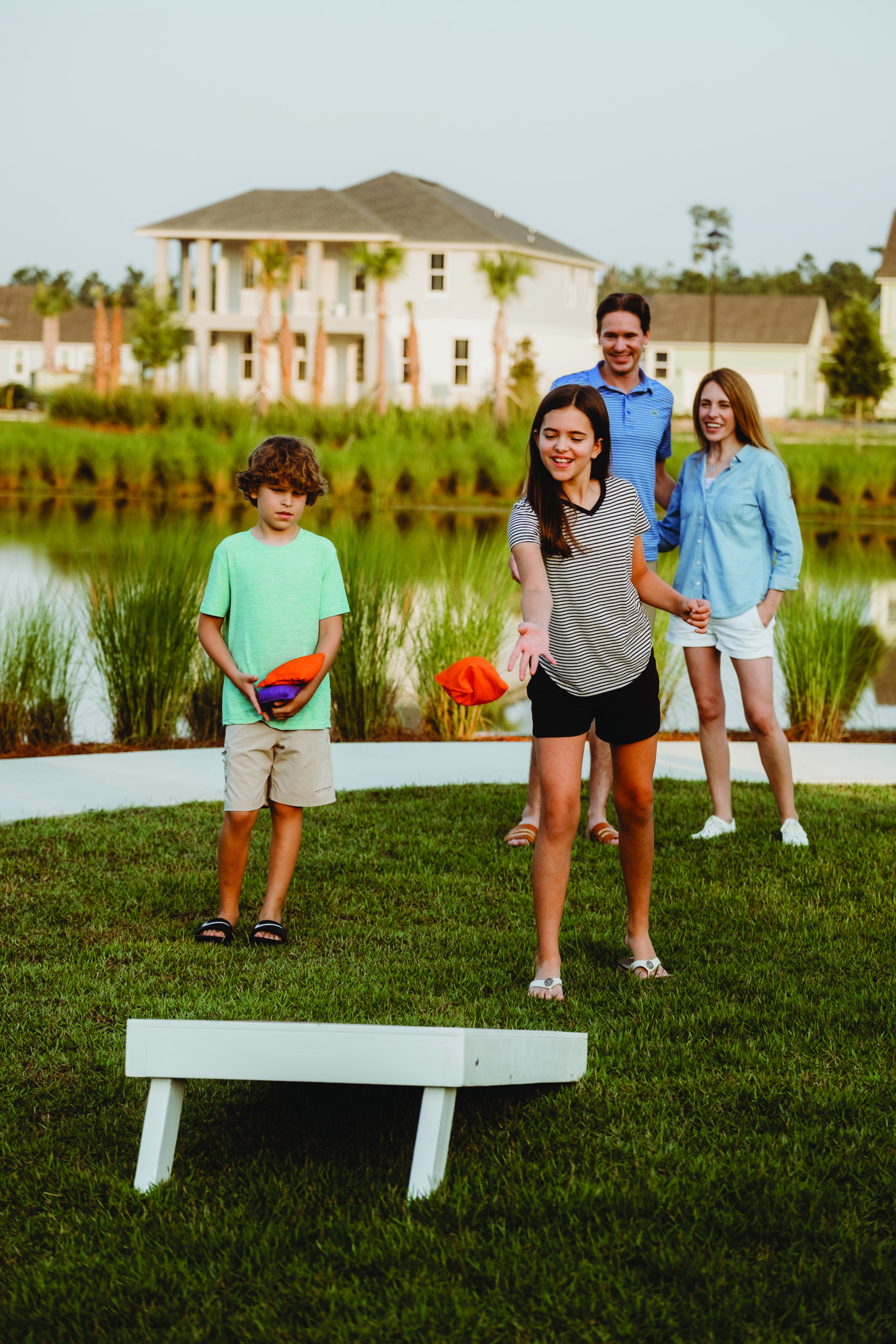 A family of four playing cornhole. The girl is about to throw her bean bag as the family watches. They are standing in a grassy park near a lake at Wildlight.