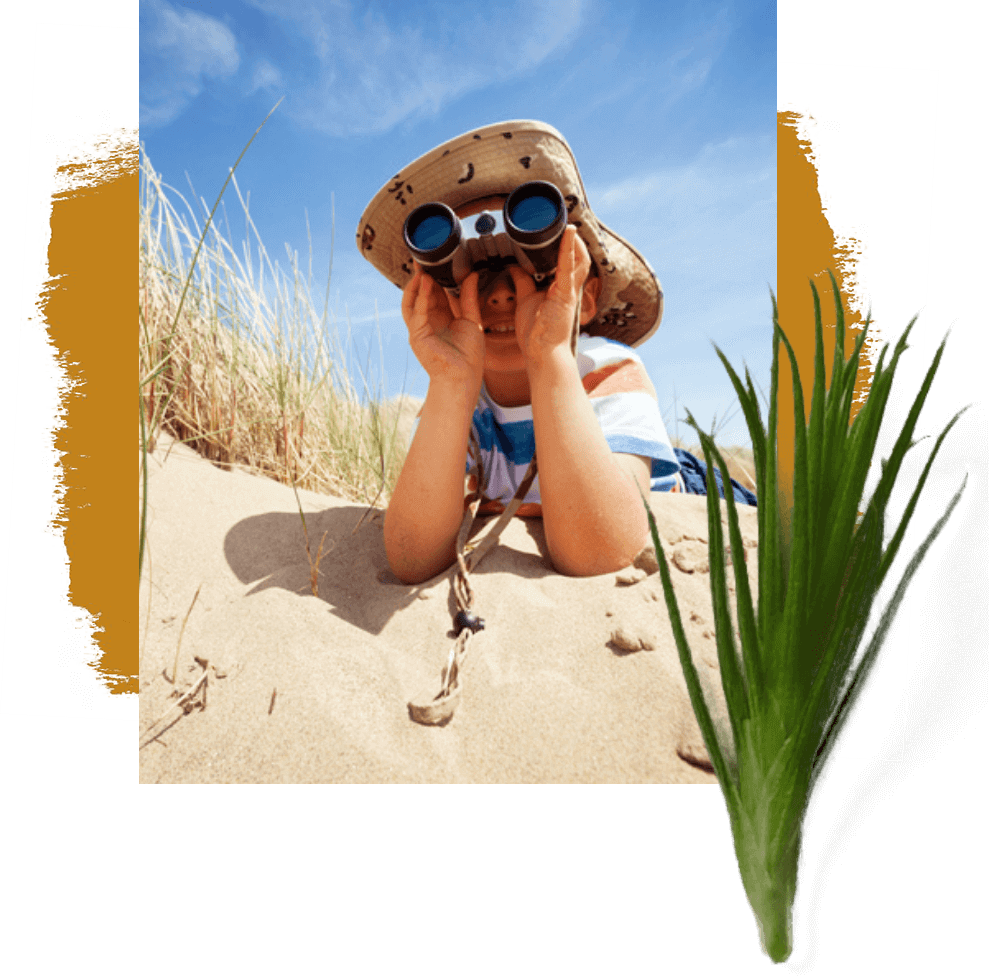 A boy in a hat looking through binoculars in the sand.