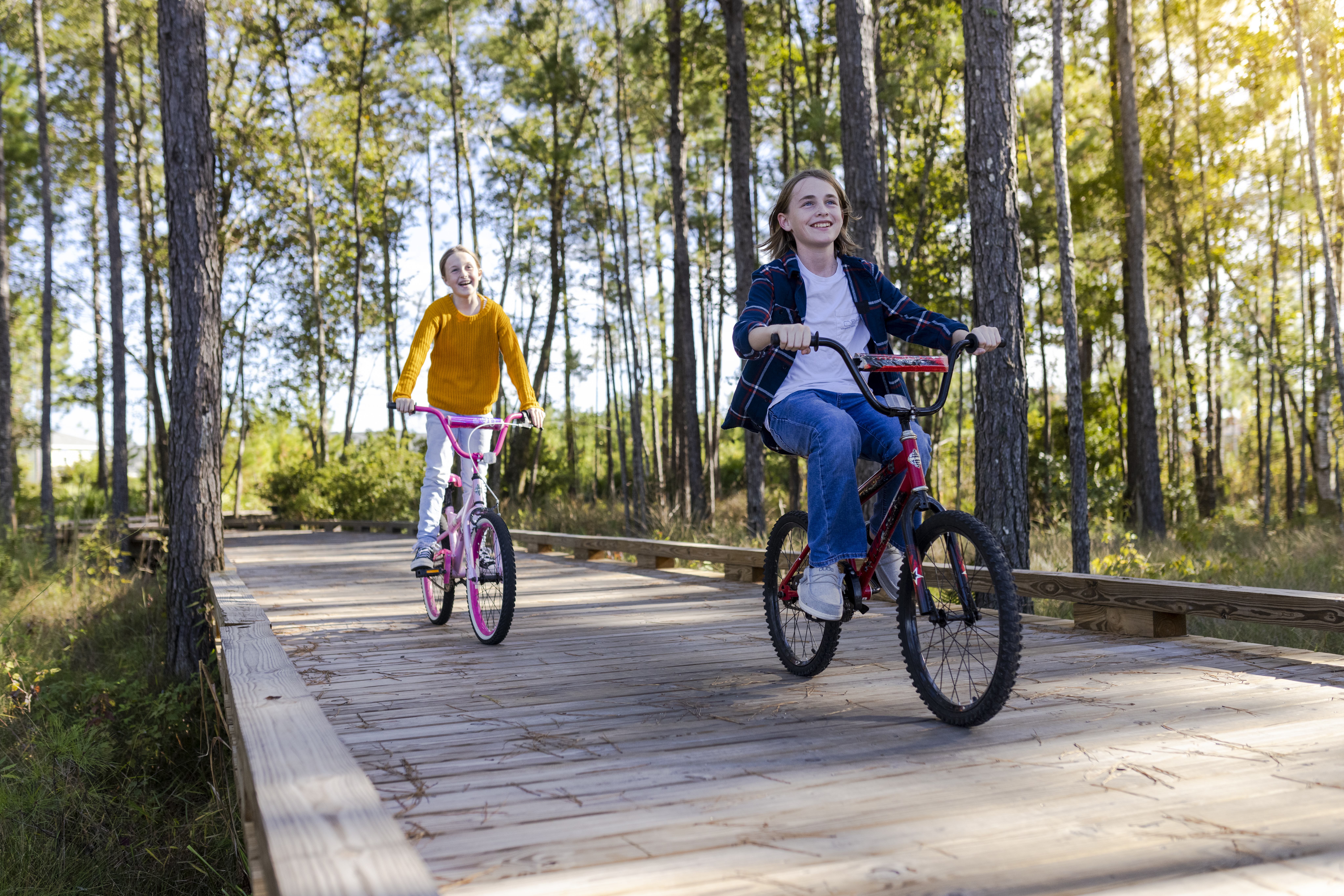 Two children ride bicycles down a boardwalk path in the woods.