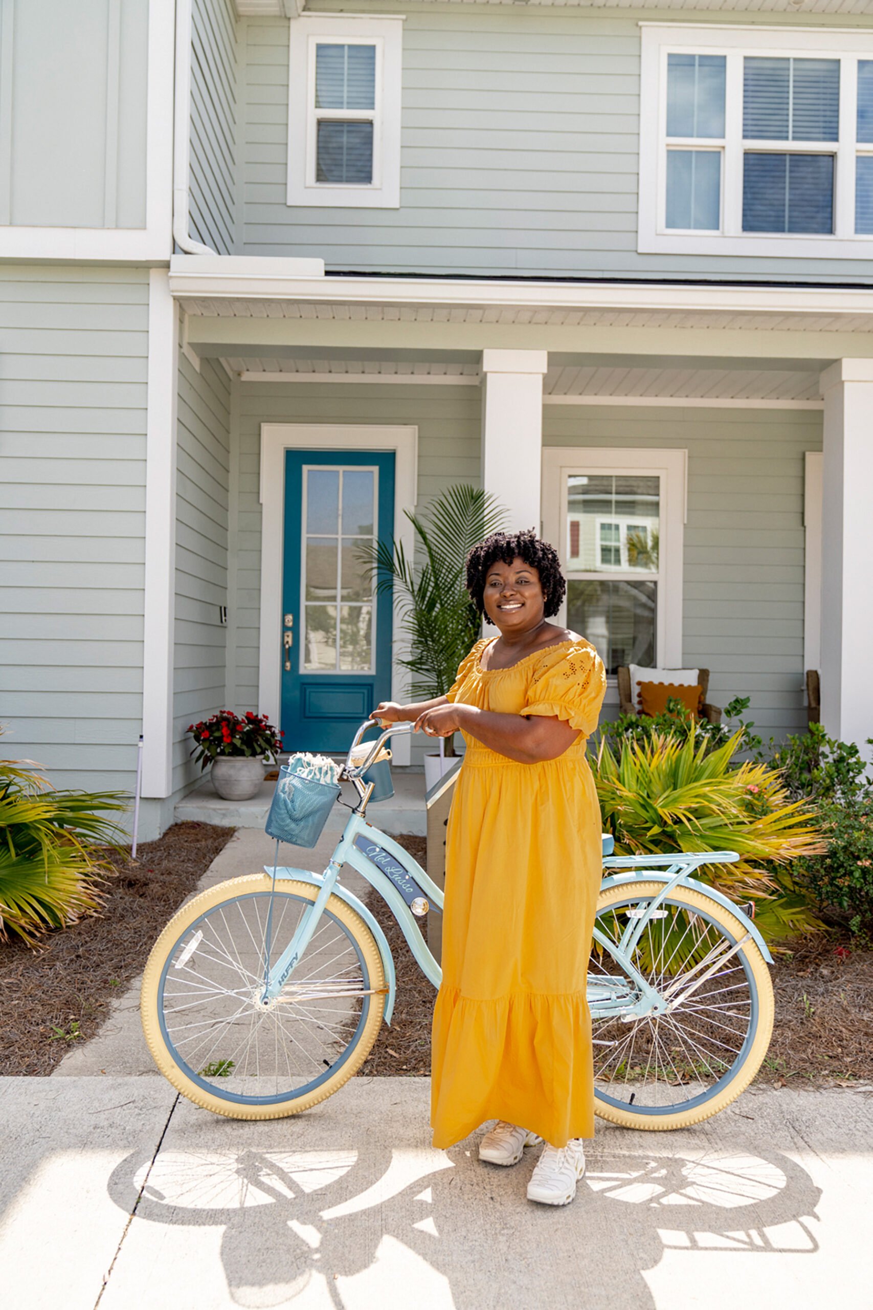 A woman in a yellow dress standing next to a blue bicycle in front of a house.