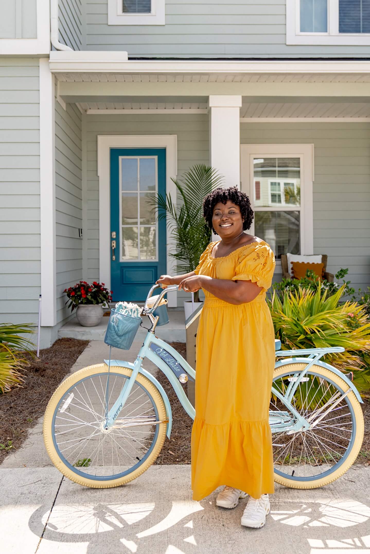A woman in a yellow dress standing in front of a house with a blue bicycle.