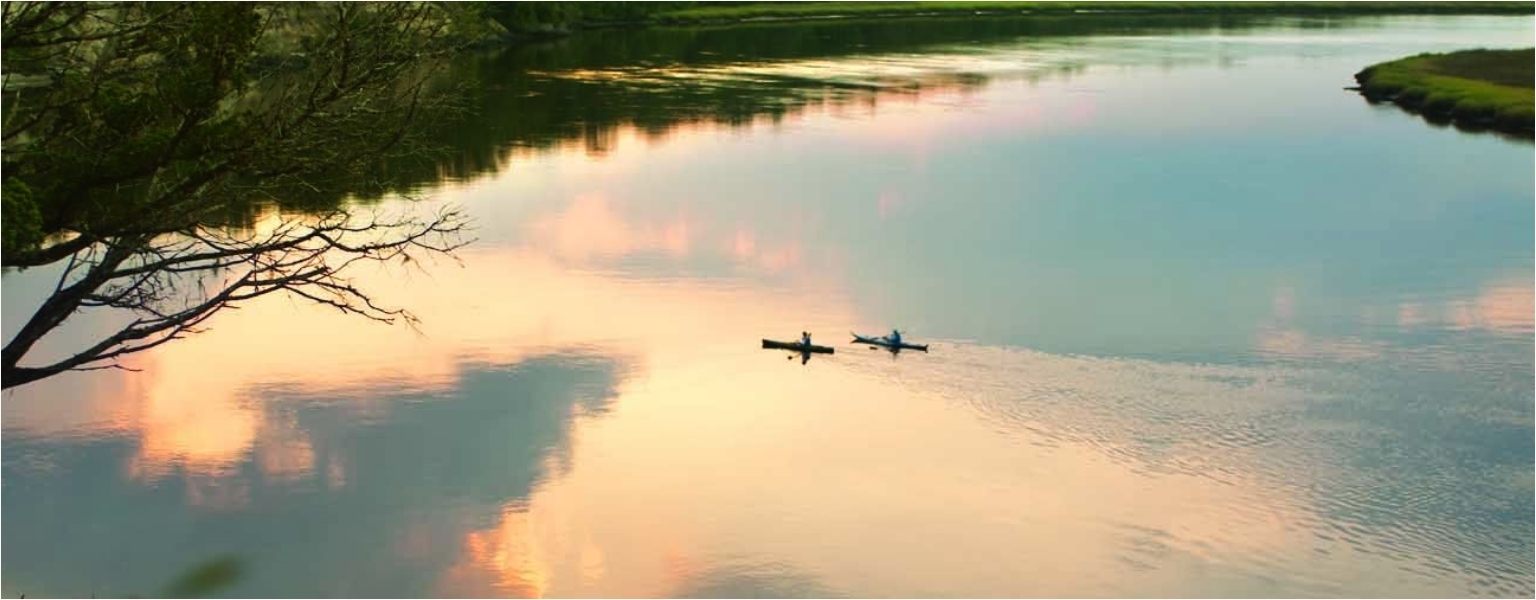 Two people paddle kayaks on a calm river at sunset, with trees and clouds reflected on the water’s surface.