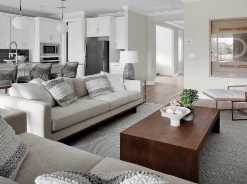 Modern living room with beige sofas, gray and white pillows, a wooden coffee table, indoor plants, and a view of the kitchen—perfect for new homes in Wildlight FL within the premier Wildlight master planned community.