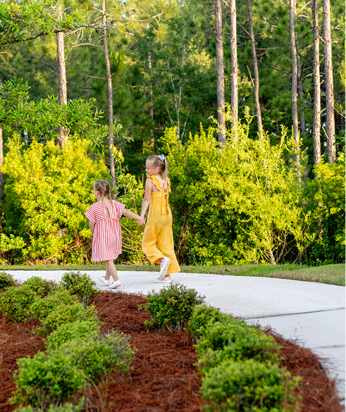 Two young girls, one in a pink striped dress and the other in a yellow outfit, walk hand-in-hand on a sidewalk surrounded by greenery and trees in the welcoming Wildlight Yulee Florida community.