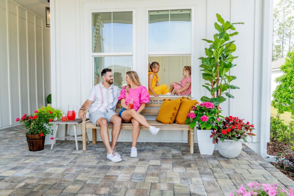 A man and woman sit on a bench with drinks on a porch in the Wildlight Yulee Florida community, while two children talk through an open window behind them. The area is decorated with plants and flowers.