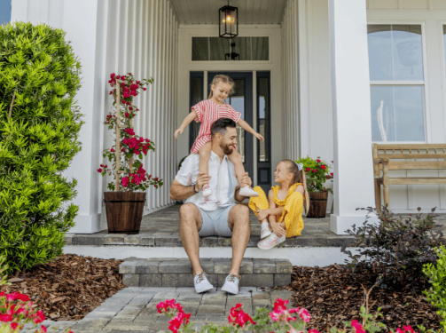 A man sits on porch steps with two young girls outside a white house, surrounded by flowers and greenery in the Wildlight Yulee Florida community.