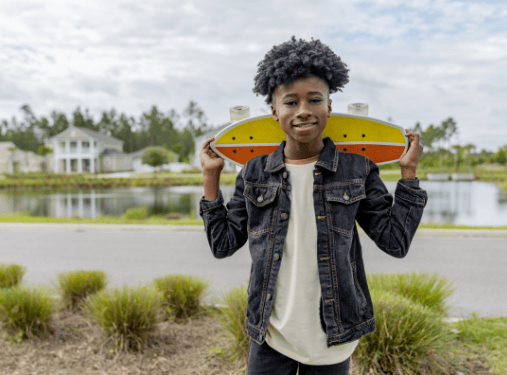 A smiling young person stands outdoors with a skateboard behind their shoulders in the vibrant Wildlight Yulee Florida community, surrounded by houses, water, and lush greenery in the background.