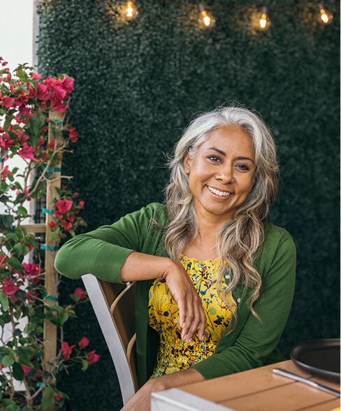 Woman with long gray hair, wearing a yellow floral dress and green cardigan, sits at an outdoor table surrounded by greenery and pink flowers, capturing the vibrant lifestyle of Wildlight Florida.