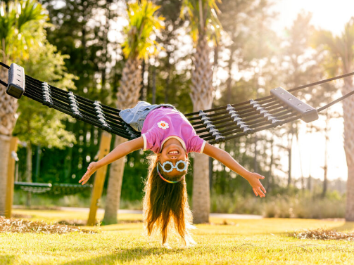 A young girl wearing sunglasses and a pink shirt hangs upside down on a black hammock outdoors, with trees and sunlight in the background—capturing the playful spirit of families living in the Wildlight master planned community.