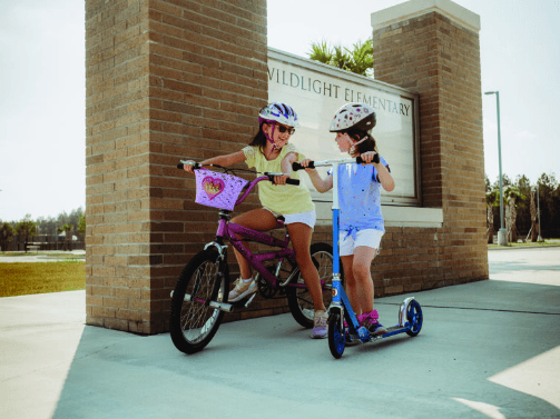 Two young girls wearing helmets smile and interact; one is on a bike and the other is on a scooter in front of a sign that reads "Wildlight Elementary" in the vibrant Wildlight master planned community of Nassau County, Florida.