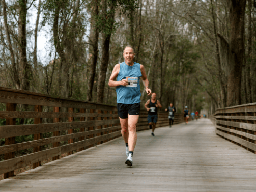 A man runs on a wooden boardwalk through a forested area in Wildlight Florida, wearing a blue tank top and shorts, with other runners visible in the background.