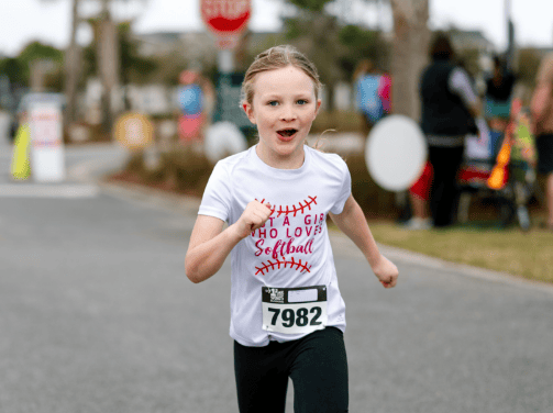 Young girl wearing a race bib runs outdoors during a race event in Wildlight Florida, smiling and sporting a white "Softball" T-shirt—capturing the lively spirit of the Wildlight master planned community.