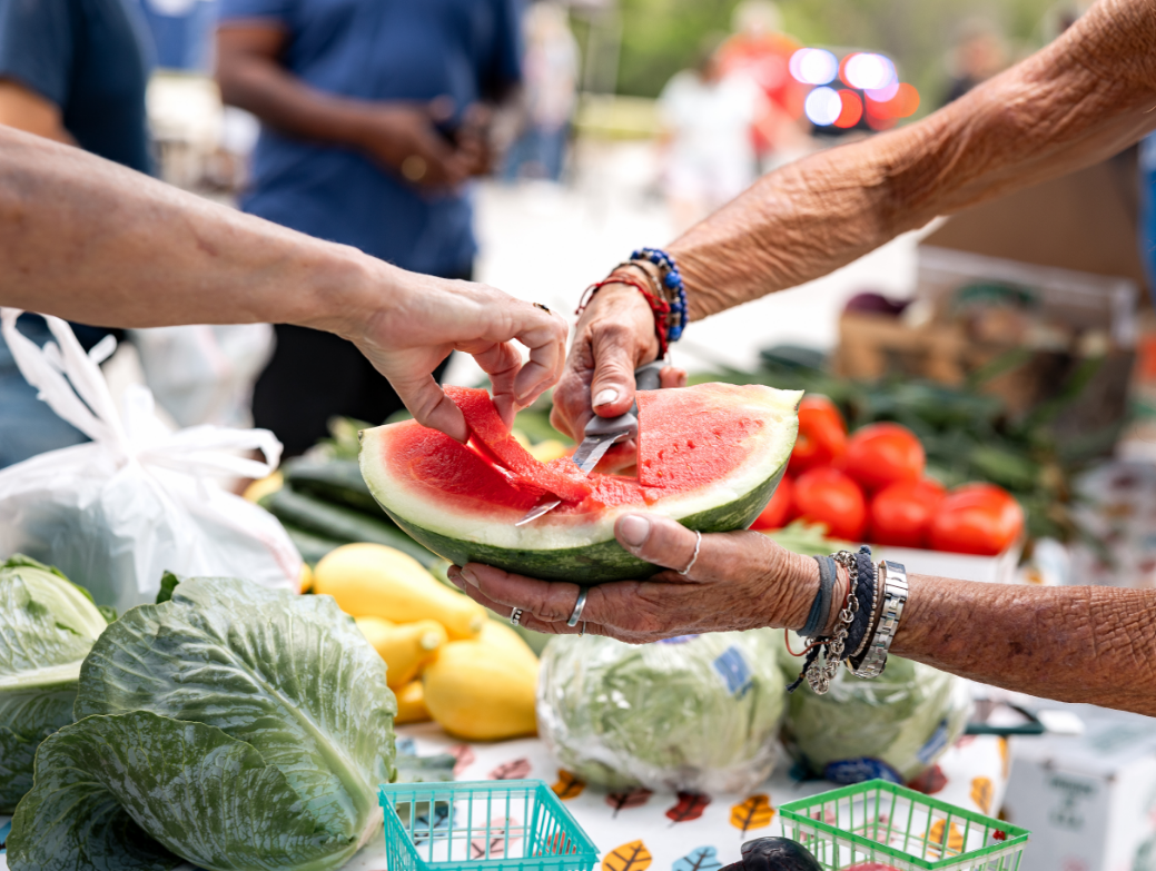 Two people cut and share a slice of watermelon at an outdoor market stand with fresh vegetables, savoring the vibrant lifestyle found in new homes in Wildlight FL, part of the welcoming Wildlight master planned community.
