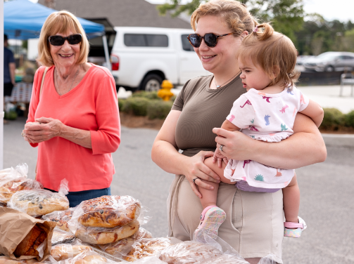 Two women and a young child stand at an outdoor market table with baked goods displayed in plastic wrap, enjoying the vibrant atmosphere of the Wildlight Yulee Florida community.