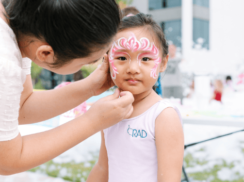 An adult paints pink and white designs on a young girl's face at an outdoor event in Wildlight Florida. The girl, wearing a white "RAD" leotard, enjoys the fun community spirit often found near new homes in Wildlight FL.