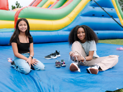 Two girls sit on a blue mat in front of an inflatable slide in the Wildlight Yulee Florida community; one ties her shoes while the other smiles at the camera. Several pairs of shoes are placed nearby.