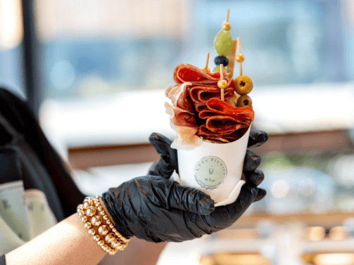 A hand in a black glove holds a cup filled with skewered cured meats, cheese, and olives—an elegant snack perfect for gatherings in the beautiful homes in Nassau County Florida.