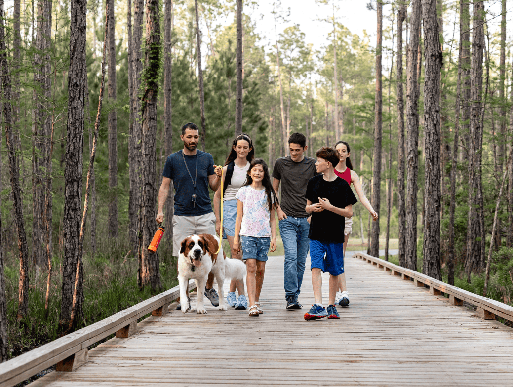 A group of people, including children and a dog, walk together on a wooden path through a forested area in the Wildlight master planned community, highlighting the nature surrounding new homes in Wildlight FL.