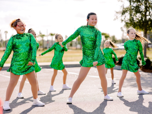 A group of girls in matching green sequin dresses and white sneakers perform a dance routine outdoors on pavement, bringing vibrant energy to Wildlight Florida—a lively community known for beautiful new homes in Wildlight FL.