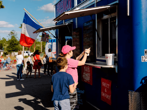 A woman and a boy stand at a blue food truck adorned with a French flag, ordering food. Nearby, the Wildlight Yulee Florida community gathers outdoors on a sunny day, with others seated and enjoying the vibrant local atmosphere.