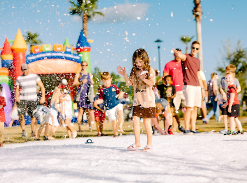 Children play in artificial snow at an outdoor event in the Wildlight Yulee Florida community, with adults and inflatable structures in the background on a sunny day.
