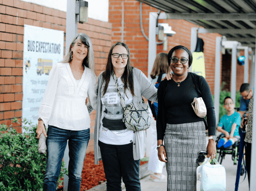 Three women stand together outside a brick building in Wildlight Florida, smiling at the camera. Other people and a sign reading "Bus Expectations" are visible in the background, capturing the spirit of community near new homes in Wildlight FL.