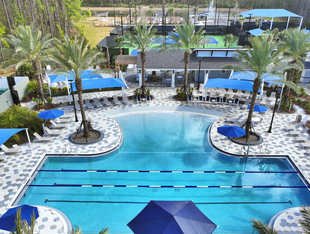 A large outdoor swimming pool with lane markers, surrounded by lounge chairs, palm trees, and blue umbrellas sits at the heart of the Wildlight Yulee Florida community, with tennis and pickleball courts in the background.