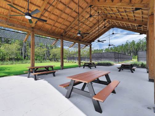 Covered outdoor pavilion with a wooden roof, ceiling fans, and picnic tables on a concrete floor. Green grass and trees are visible in the background, offering a relaxing spot in the Wildlight Yulee Florida community.