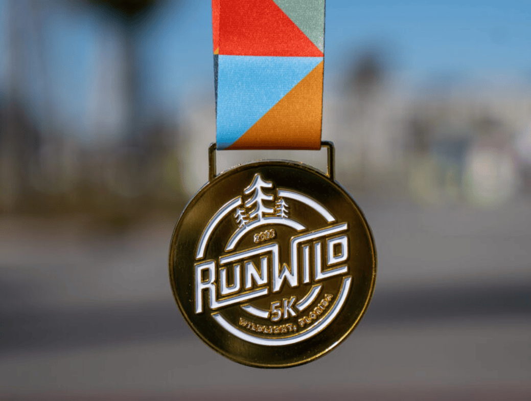 A gold "Run Wild 5K" medal hangs from a blue, orange, and red geometric-patterned ribbon against a blurred outdoor background in the Wildlight master planned community.
