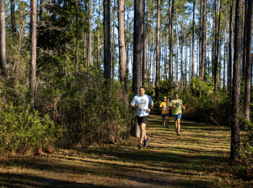 Three people are running along a dirt path through a forested area in the Wildlight Yulee Florida community, surrounded by tall trees and lush greenery.