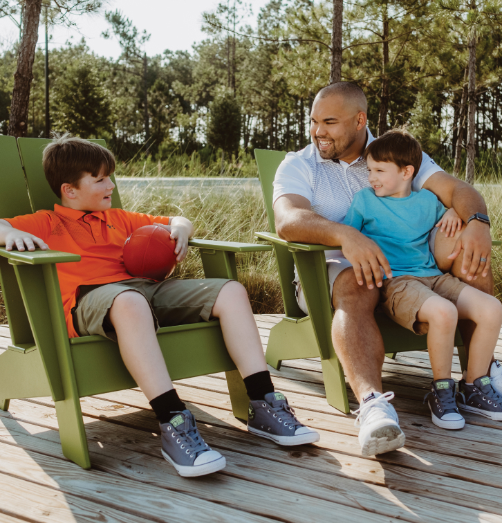 An adult and two boys sit on green chairs outdoors in the Wildlight master planned community; one boy holds a red ball, and all three are smiling and talking on a wooden deck surrounded by trees.