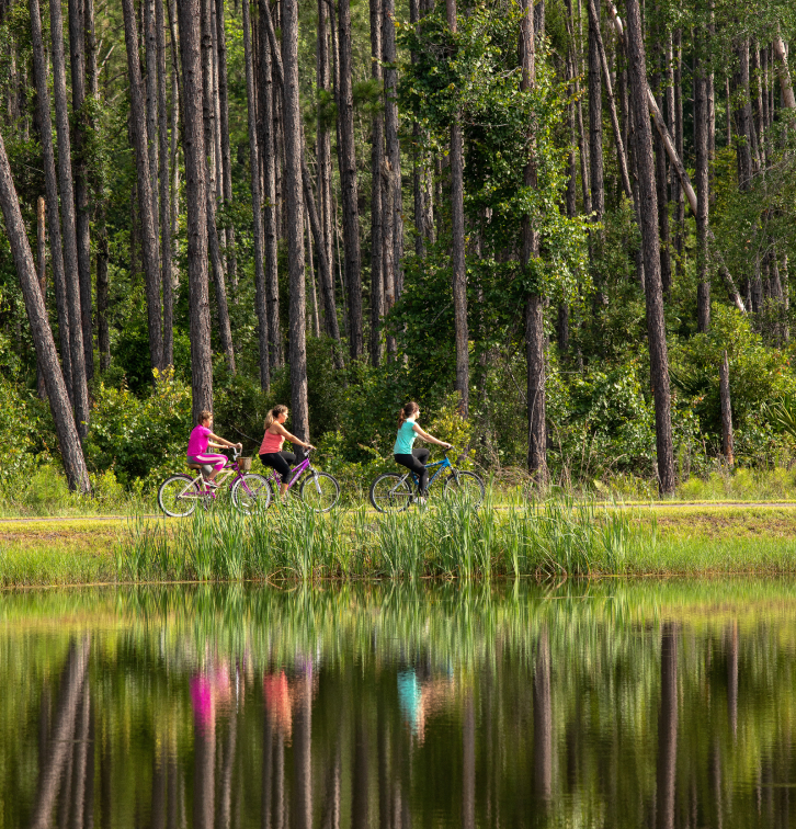 Three people ride bicycles on a path beside a forested area in the Wildlight Yulee Florida community, with their reflections visible in a calm pond in the foreground.