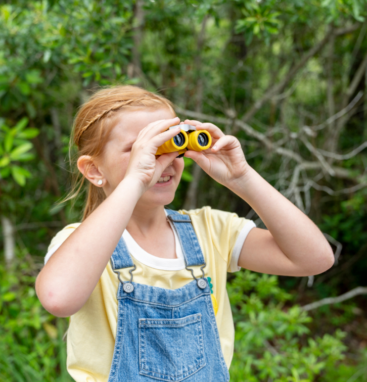 A young girl in a yellow shirt and denim overalls uses yellow binoculars to look into the distance outdoors, surrounded by green foliage in the Wildlight Yulee Florida community.