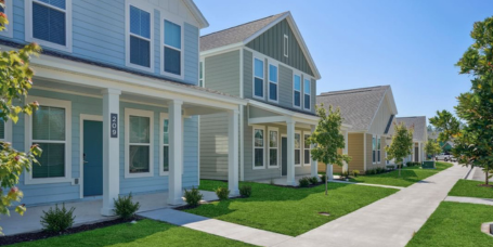 Row of modern, two-story suburban houses with front porches, green lawns, and trees line a sidewalk on a sunny day in the Wildlight master planned community.