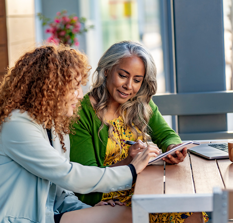 Two women sit at a wooden table in Wildlight Florida, one holding a tablet while the other writes on it. A laptop, coffee cup, and plant are visible—capturing daily life in the vibrant Wildlight master planned community.