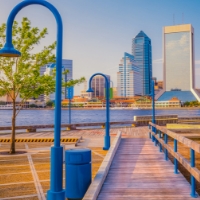 A wooden walkway with blue street lamps leads toward a river, with a city skyline in the background under a clear sky—perfect for those seeking homes in Nassau County Florida and the vibrant Wildlight Yulee Florida community.