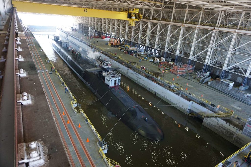 A submarine is docked in a large indoor shipyard facility within the Wildlight Florida community, surrounded by scaffolding, workers, and equipment along the sides of a narrow water channel.